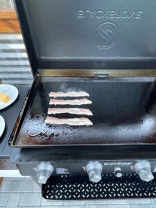 Slices of bacon sizzling on the hot Blackstone griddle, an essential ingredient in the Blackstone smash burger recipe.