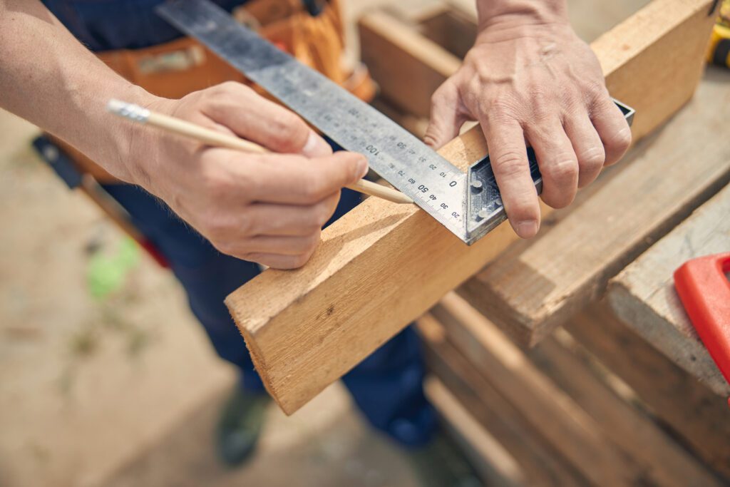 Carpenter carefully measuring the length of a wooden plank with a framing square.